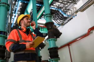 A Pronto Plumbing team member in safety gear uses a tablet and radio beside large indoor pipes, ensuring smooth operations.
