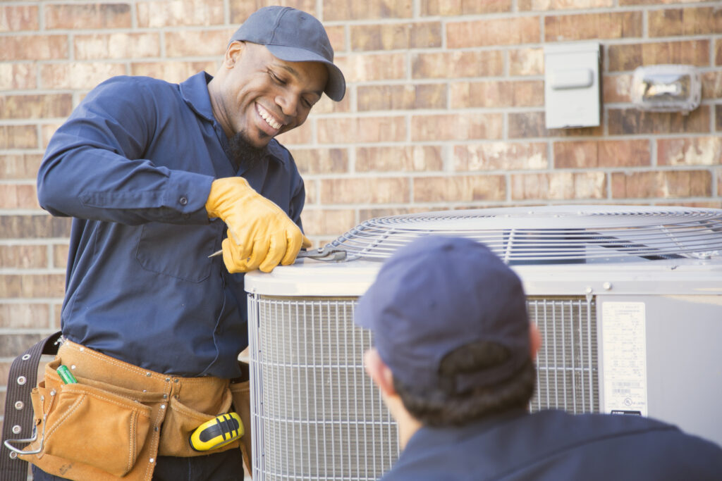 Pronto Plumbing, Heating, Air, and Drains technicians service an outdoor AC unit—one smiles with a screwdriver as the other listens.