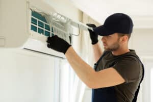 A Pronto technician in gloves and a cap inspects the filter of an indoor wall-mounted air conditioning unit for optimal performance.