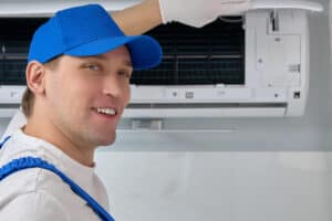 A friendly Pronto Plumbing, Heating, Air, and Drains technician inspects a wall-mounted AC unit, smiling in uniform at the camera.