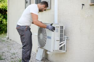 A Pronto team member wearing gloves inspects an outdoor AC unit mounted on a building’s exterior wall during a service visit.