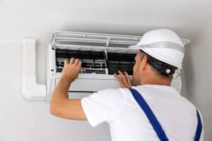 A Pronto Plumbing, Heating, Air, and Drains technician in safety gear checks and adjusts filters on a wall-mounted AC unit.