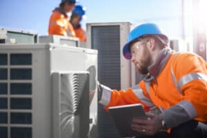 A Pronto Plumbing, Heating, Air, and Drains technician inspects an outdoor HVAC unit with a tablet as team members work nearby.