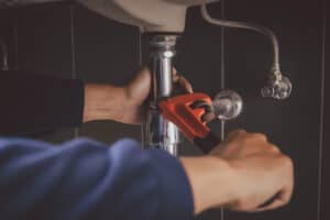 A Pronto Plumbing technician uses a red pipe wrench under a sink, expertly adjusting a pipe for quality service.