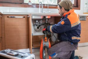 A Pronto Plumbing technician clears a kitchen sink drain using professional equipment, tools organized nearby and cabinet doors open.