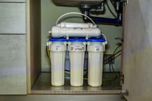 A Pronto Plumbing water filtration system with three labeled filters is neatly installed under a kitchen sink with pipes and cabinet doors visible.
