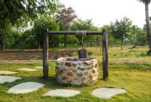 A stone water well with a wooden frame and bucket sits on grass near stepping stones and trees, perfect for Pronto Plumbing’s services.