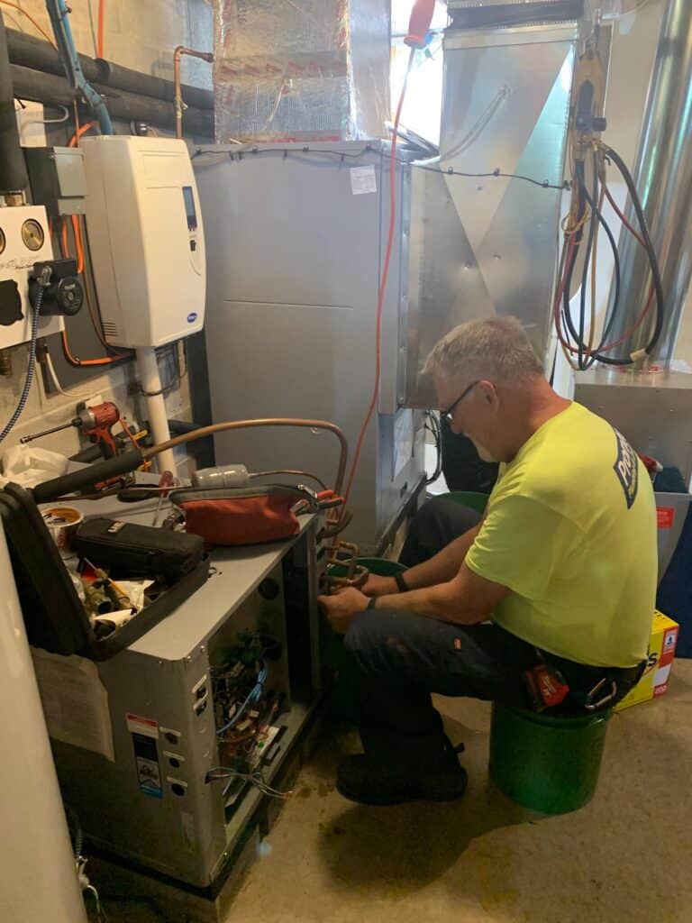 A friendly Pronto Plumbing, Heating, Air, and Drains technician, dressed in a neon yellow uniform, performs professional repairs on an HVAC unit located in a basement.